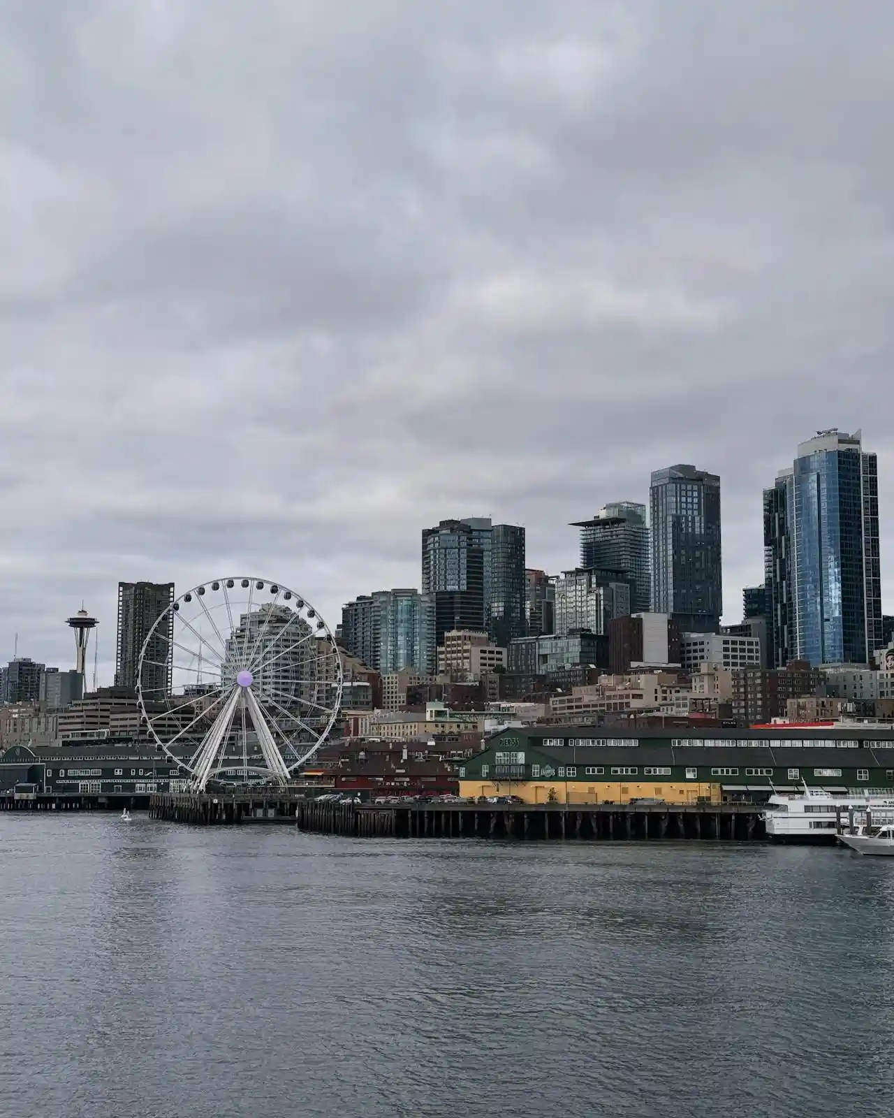 Seattle waterfront with the Great Wheel ferris wheel and downtown skyscrapers under a cloudy sky.