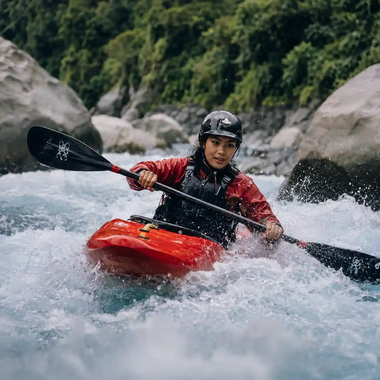 Person in red kayak paddling through white water rapids near rocks and forest.
