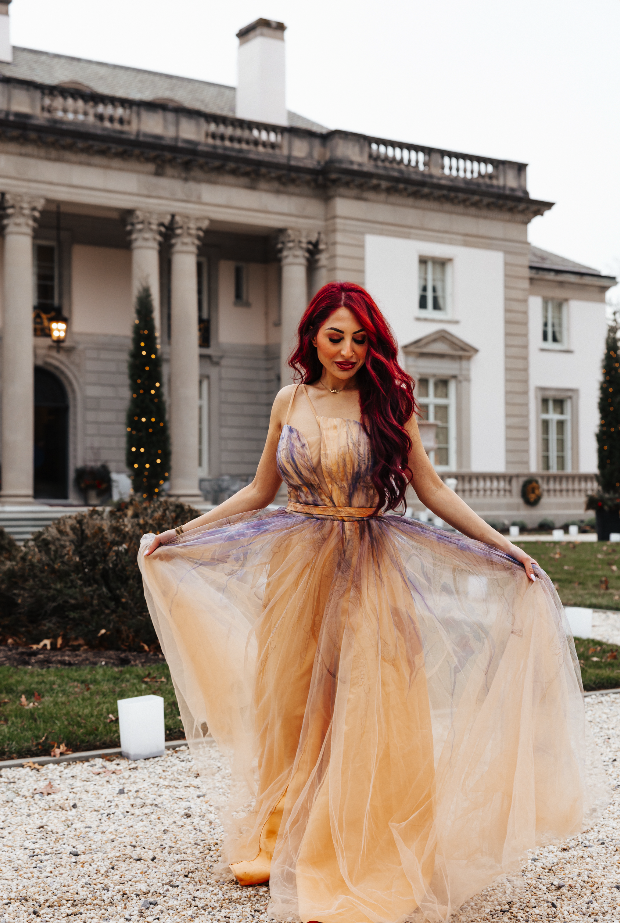 Woman with long red hair in a flowing gold and purple gown standing on a gravel path outside a mansion.