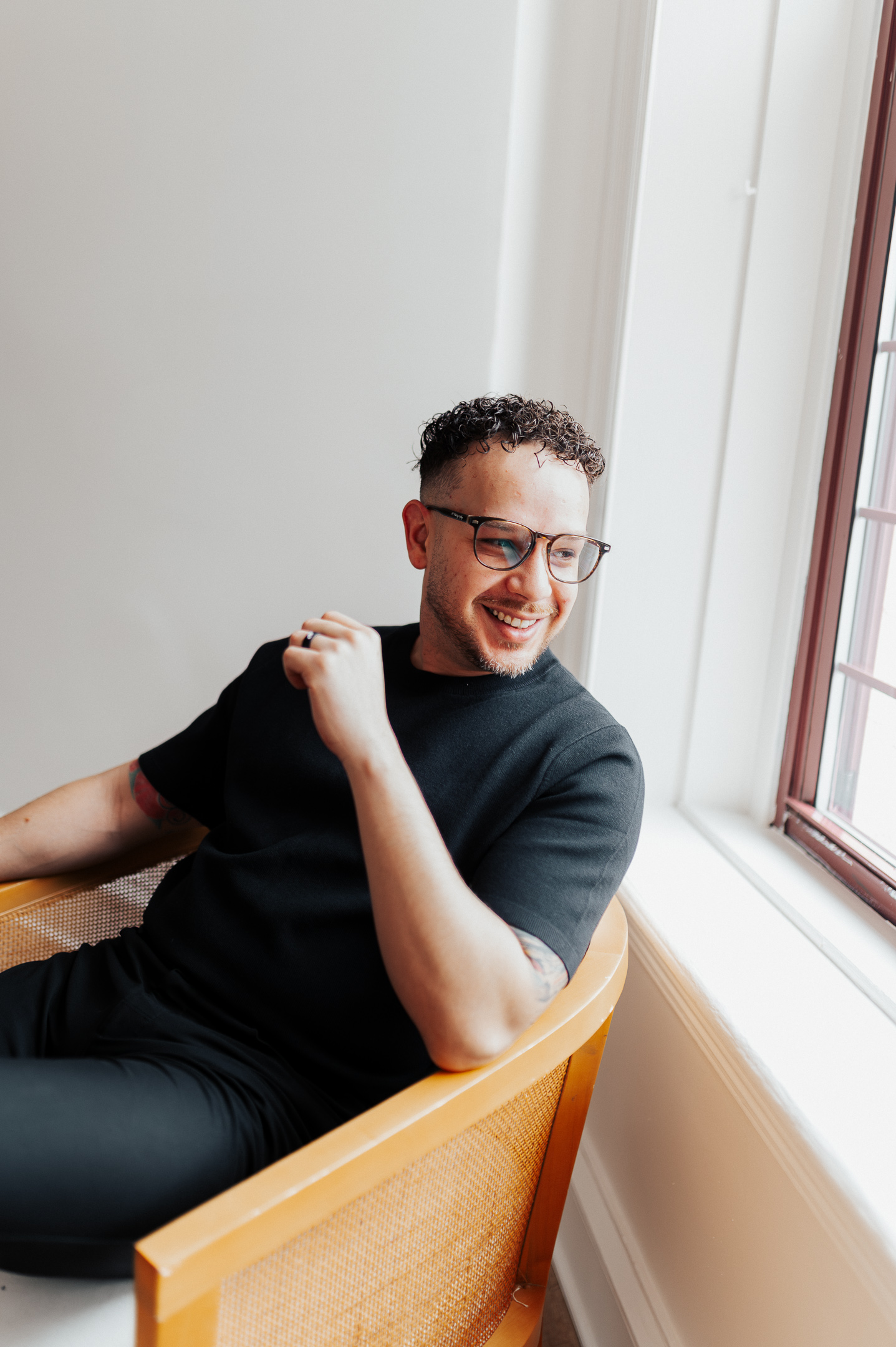 Smiling man with glasses and tattoos sitting on a wooden chair by a window in a bright room.