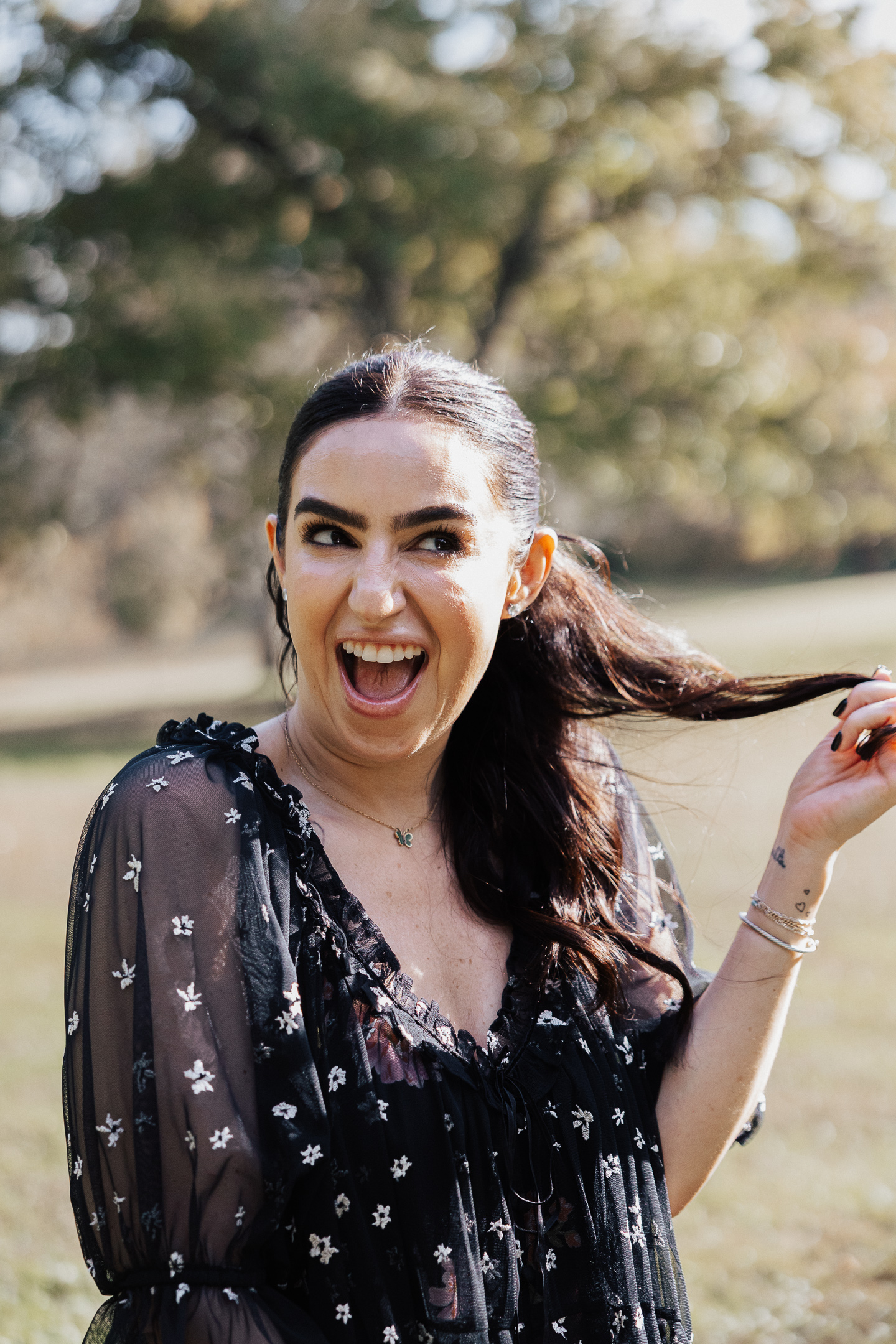 Happy woman outdoors with dark hair in a black floral dress pulling her ponytail and laughing.