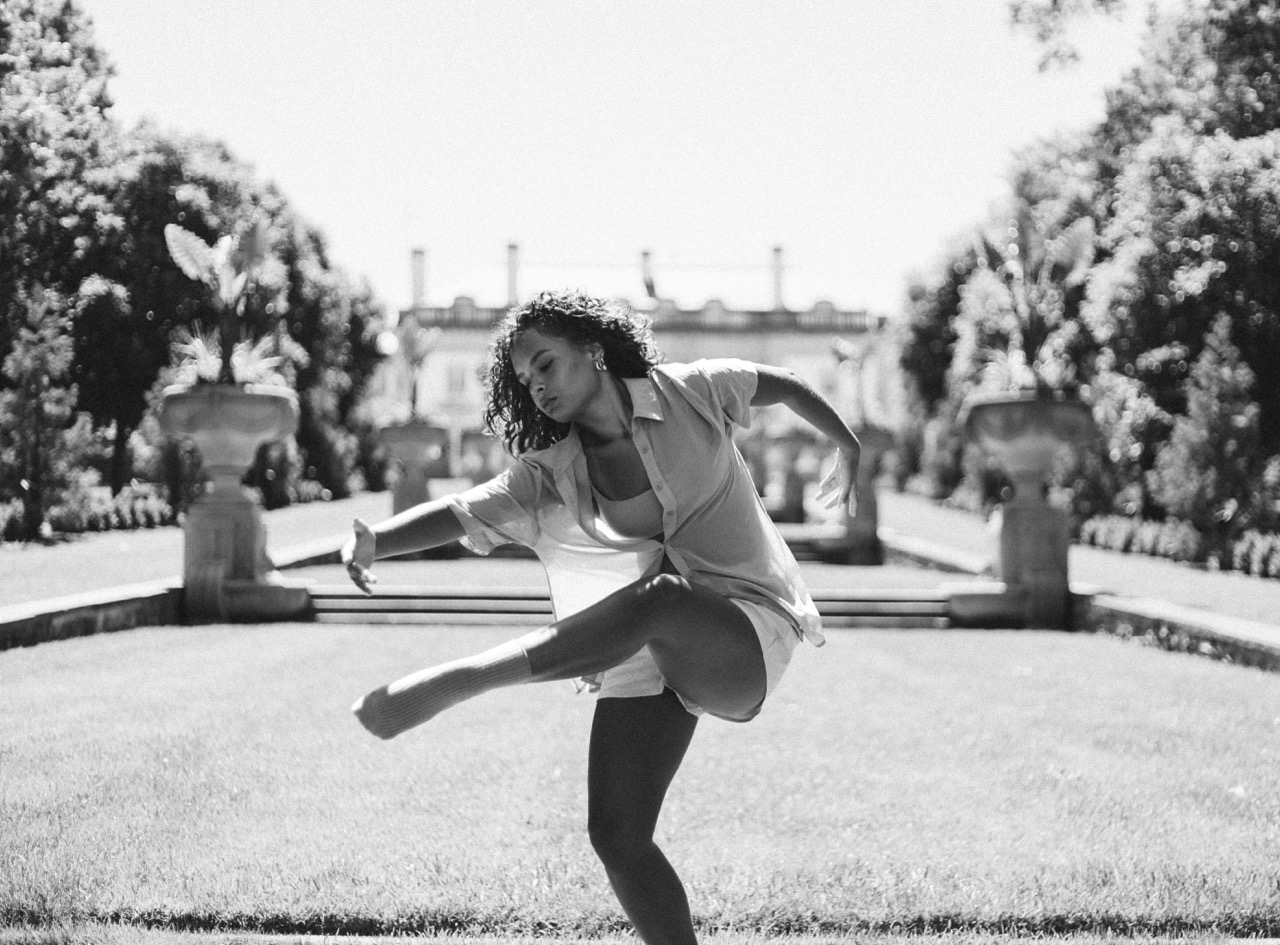 Woman in light shirt and shorts dancing outdoors in a garden with stone urns and trimmed bushes.