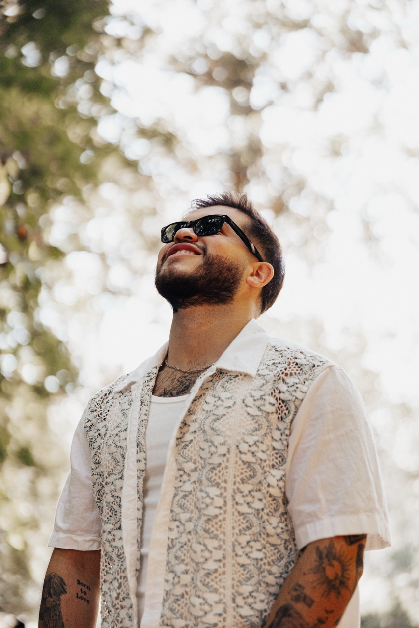 Man with beard and sunglasses wearing a patterned short-sleeve shirt looking upward outdoors.