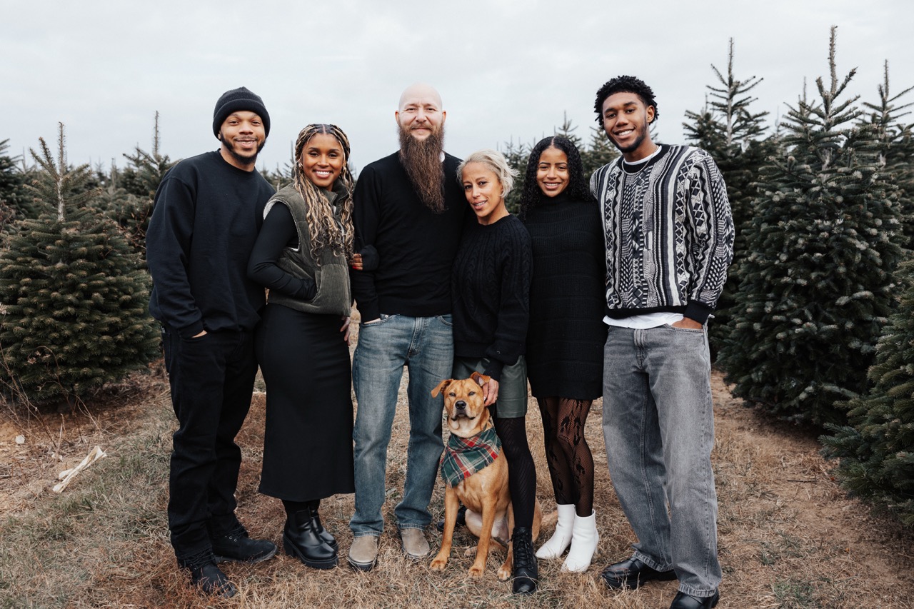 Six people and a dog standing among Christmas trees outdoors, all smiling at the camera.