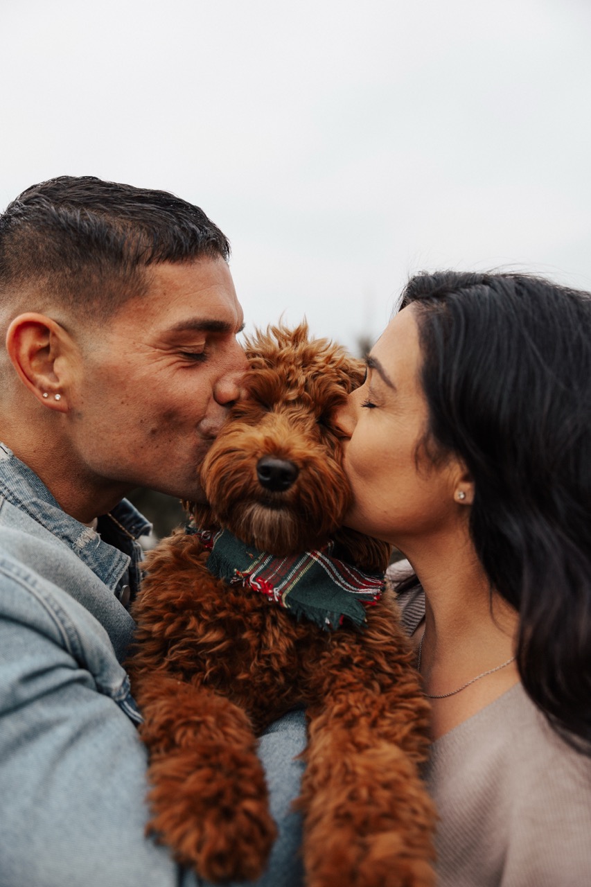 Man and woman each kissing a brown fluffy dog wearing a green plaid scarf outdoors.