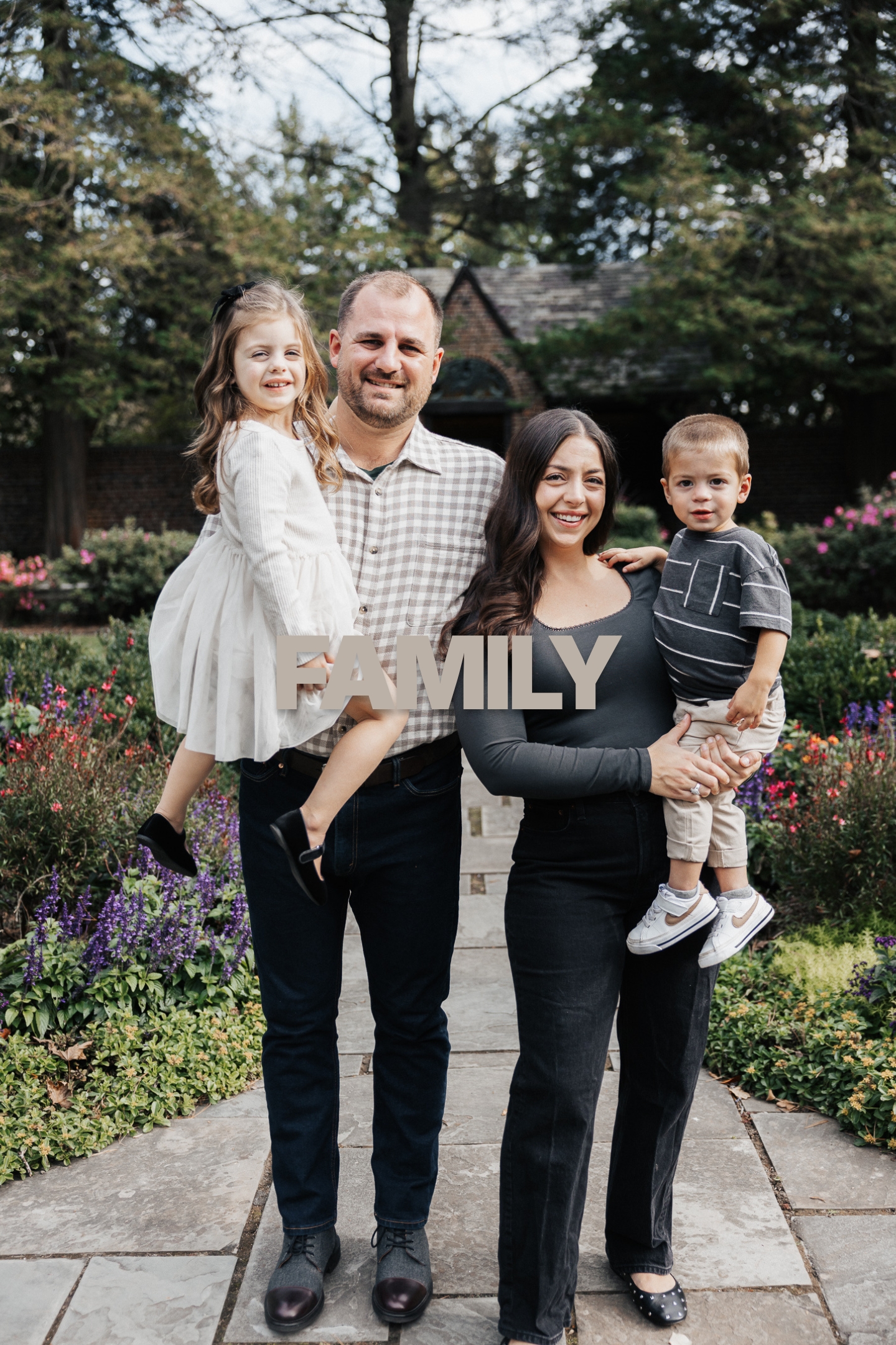 Smiling family of four standing on garden path, parents holding two young children with flowers around.