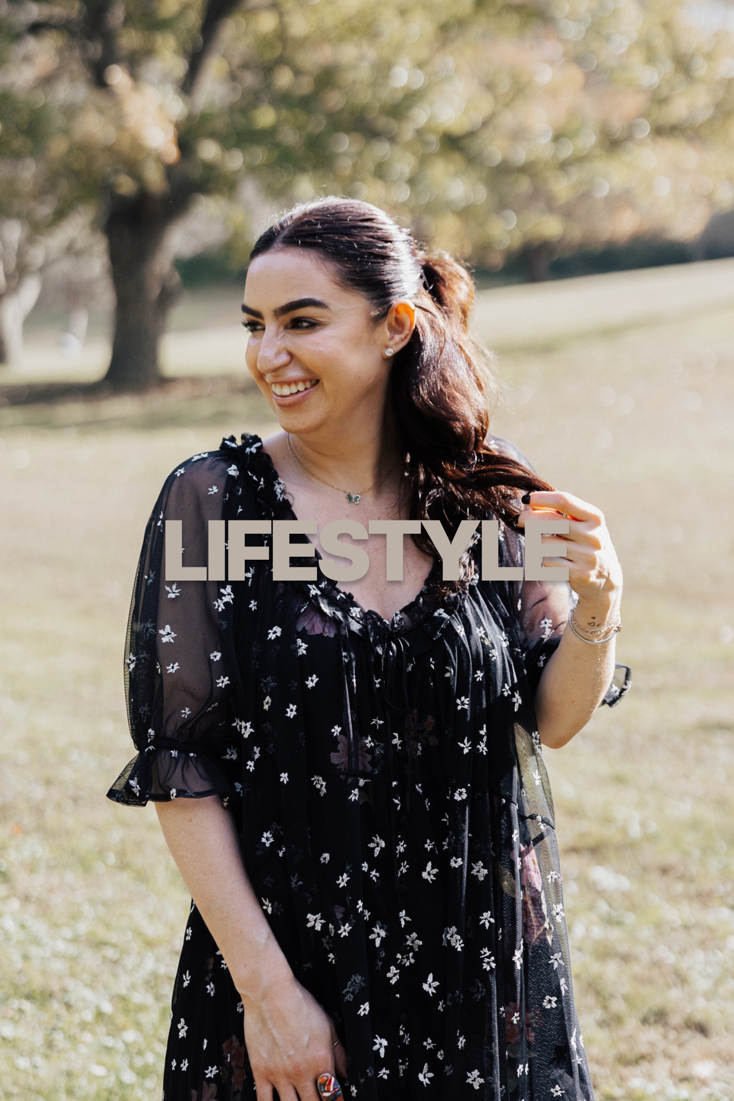 Smiling woman in black floral dress outdoors on sunlit grassy area with trees in background.