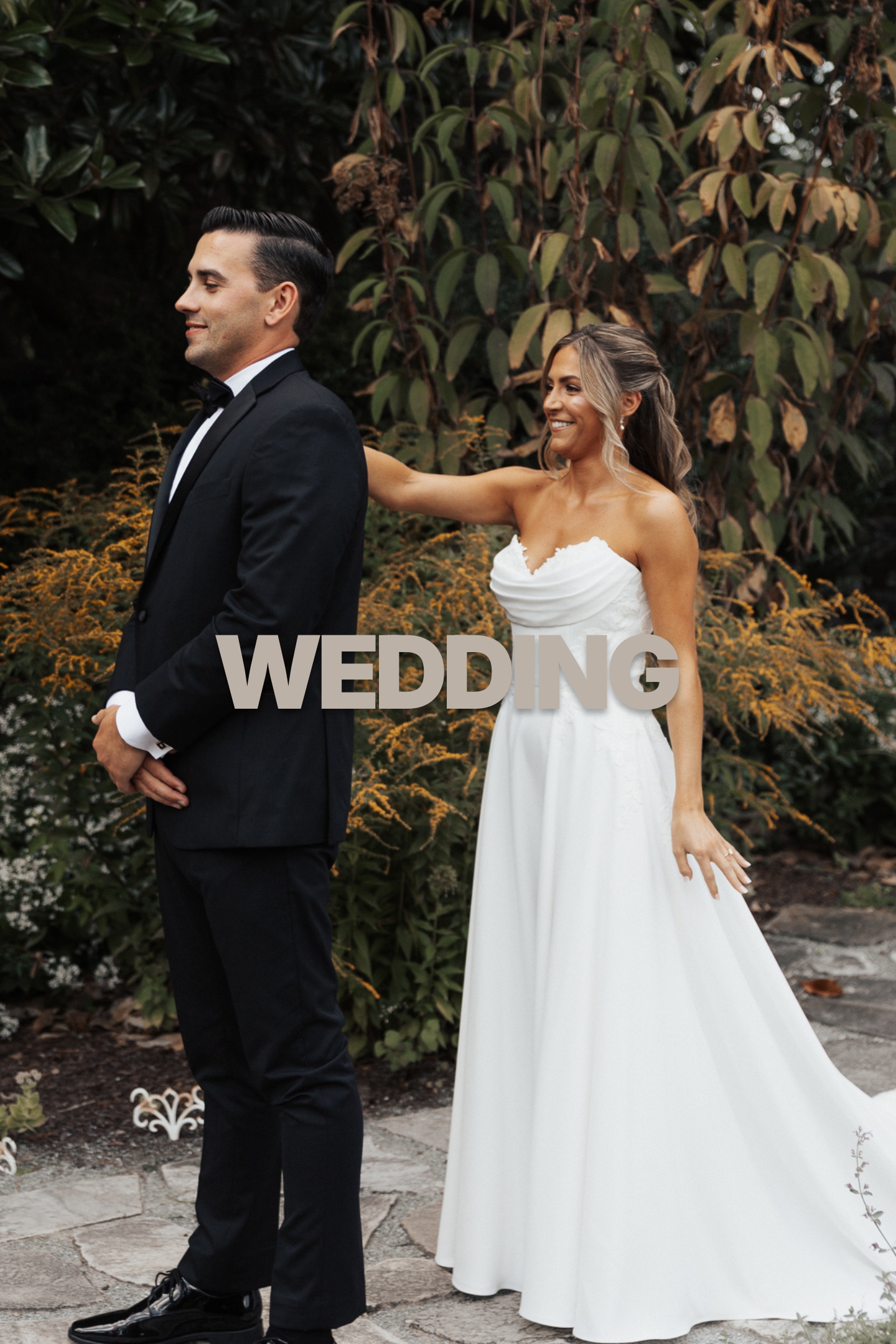 Bride smiling with hand on groom’s shoulder while groom stands back facing away in a garden.