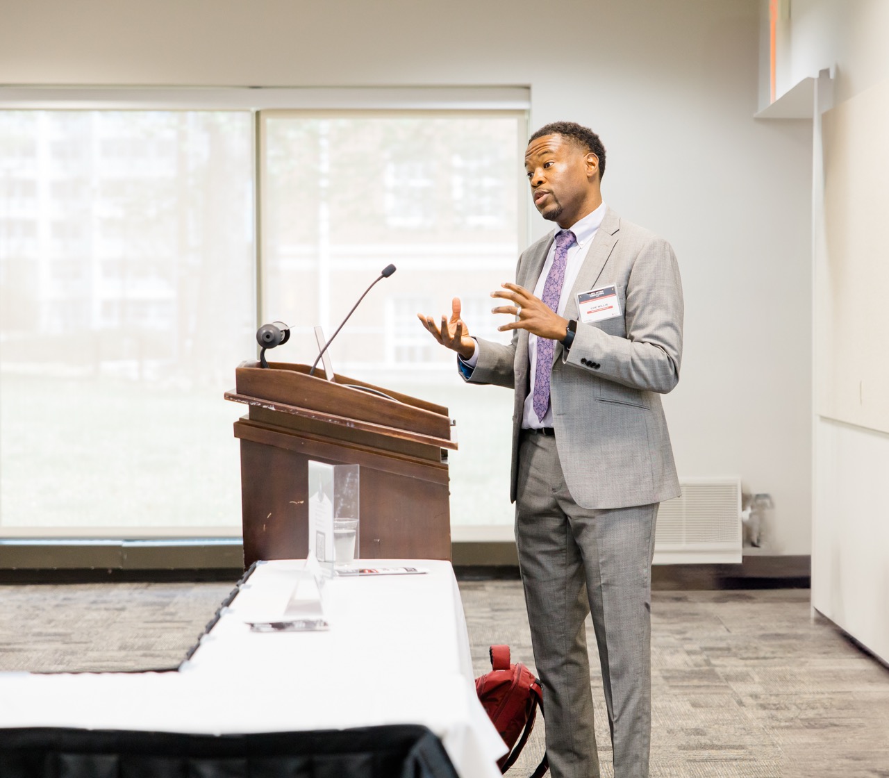 Man in gray suit and purple tie speaking at a podium in a conference room.
