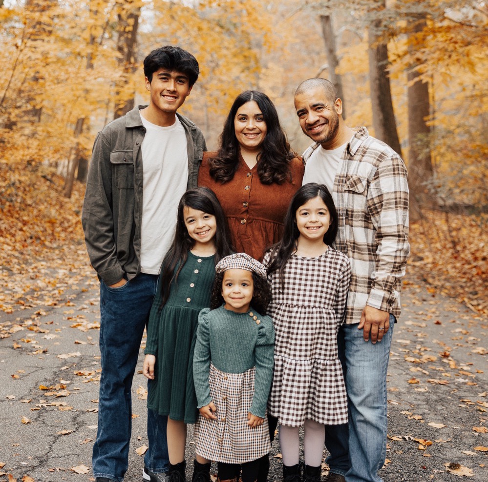 Smiling family of six posing outdoors on a leaf-covered path with autumn trees in the background.