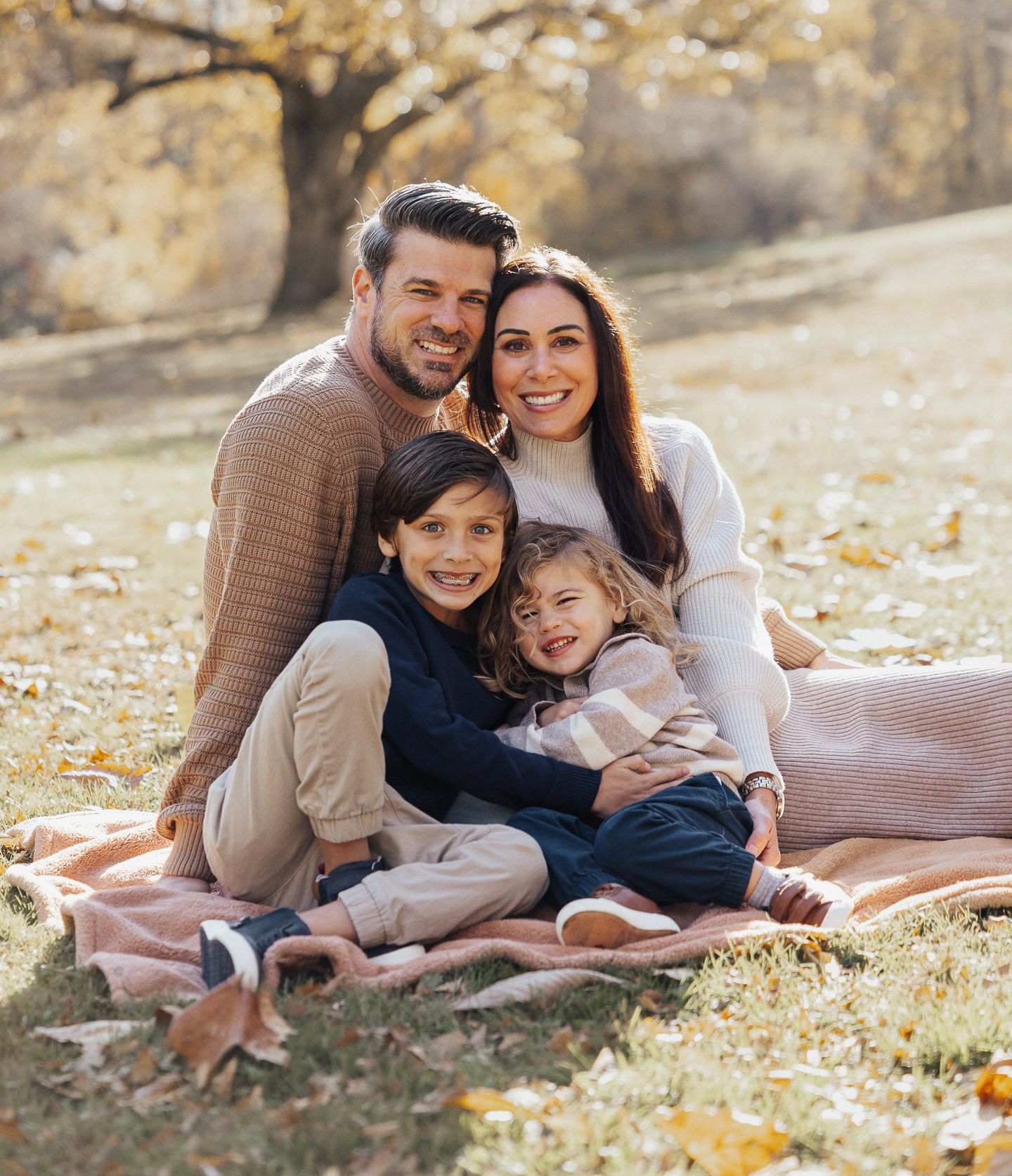 Smiling family of four sitting on blanket outdoors with autumn leaves around.