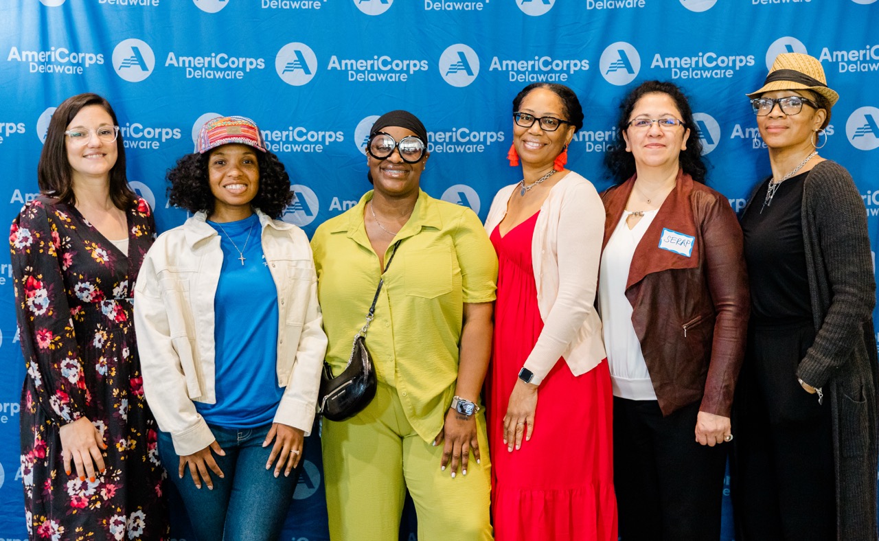 Six women standing in front of an AmeriCorps Delaware backdrop, smiling at the camera.