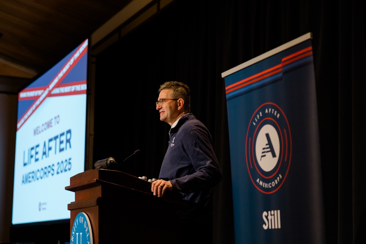 Man in glasses speaking at podium with Life After AmeriCorps 2025 screen and banner in background.