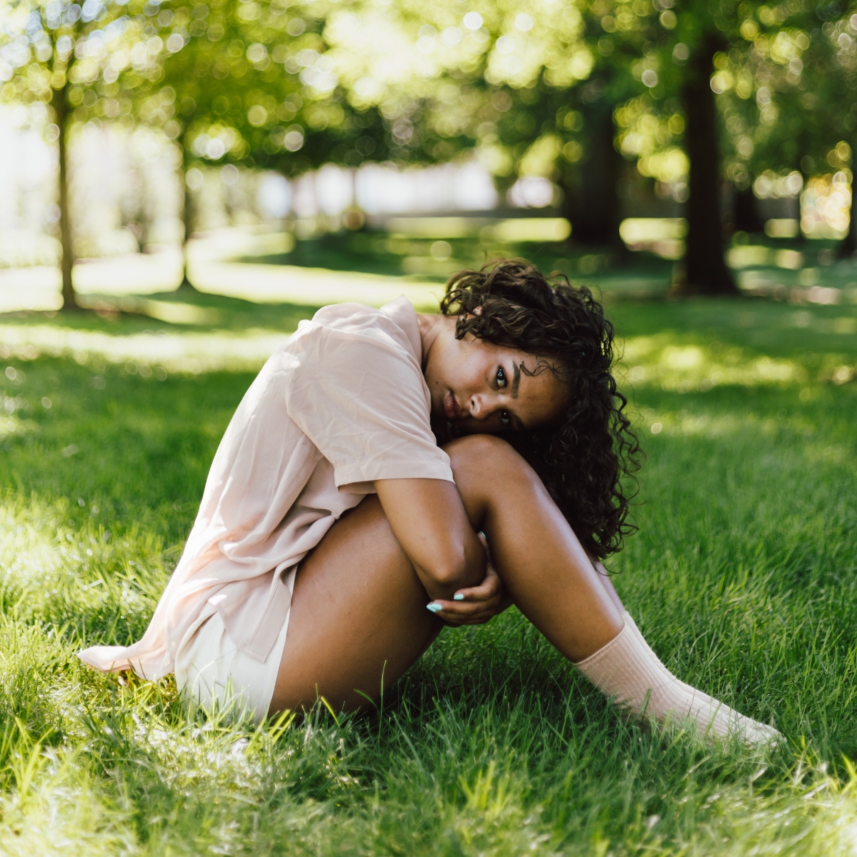 Woman with curly hair sitting curled on grass in a sunlit park with trees in the background.