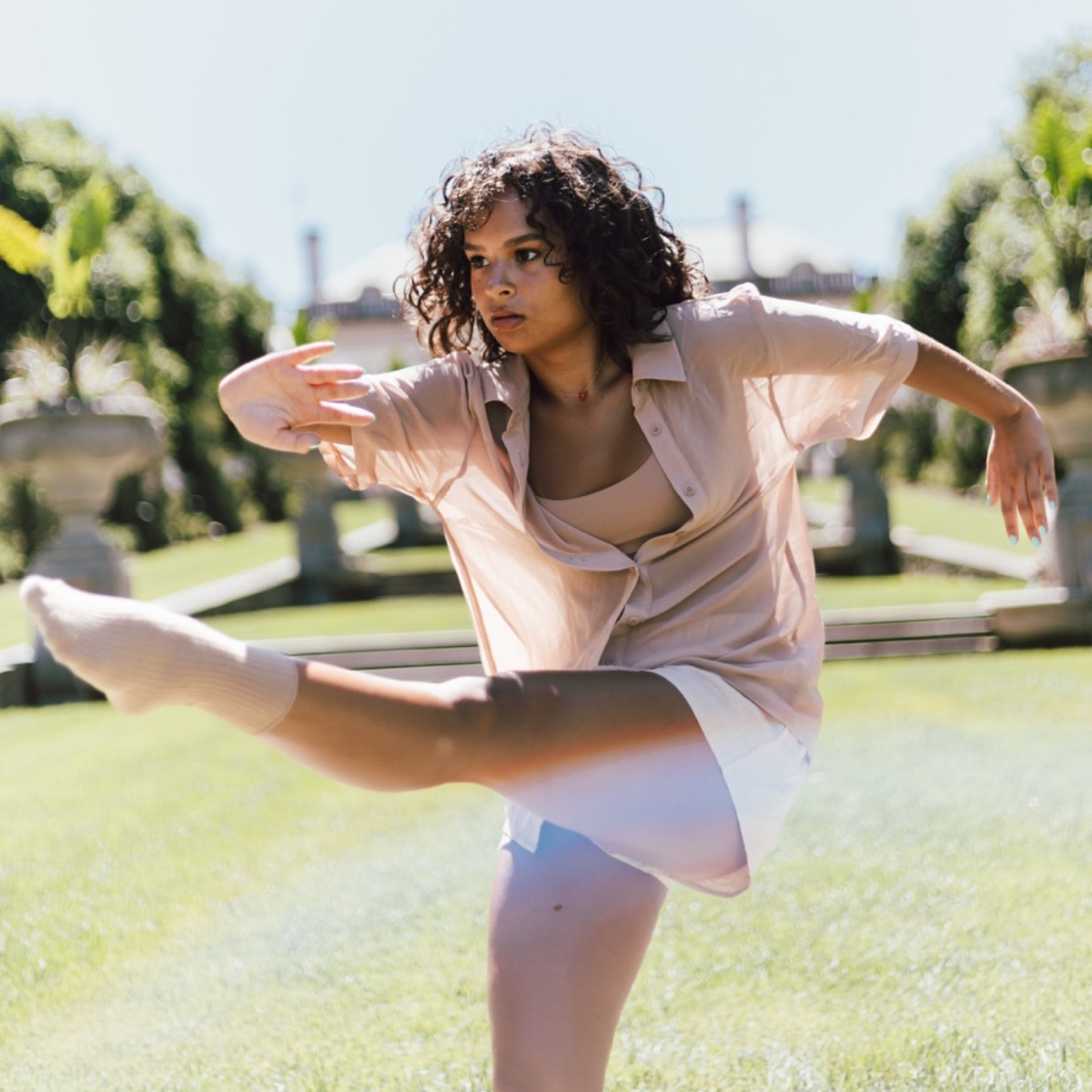 Young woman with curly hair performing a dynamic dance move outside on grass in daylight.