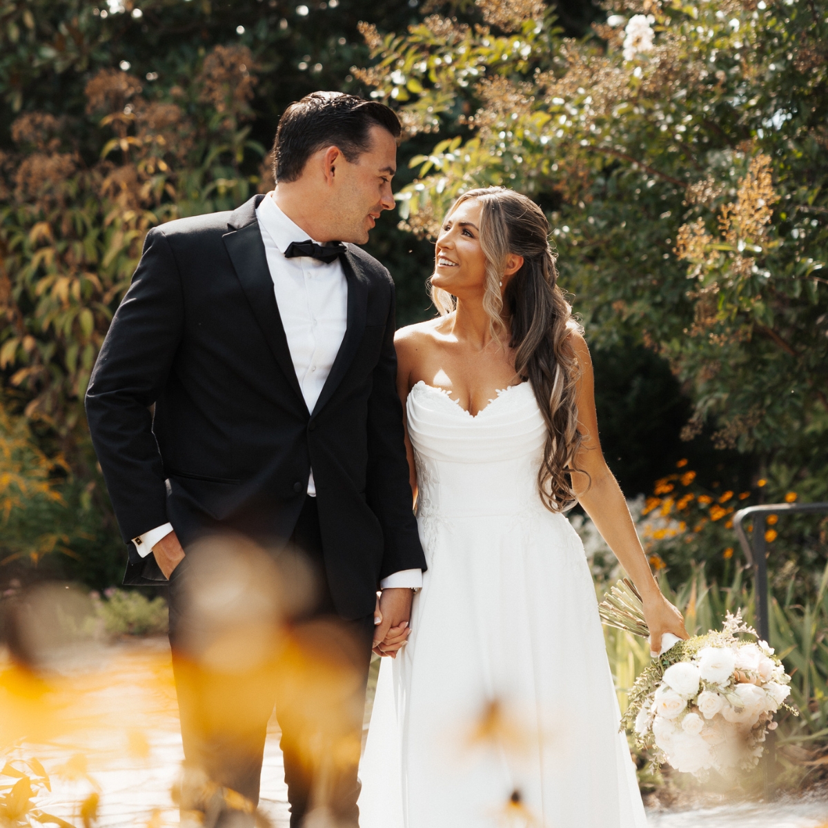 Bride and groom holding hands smiling at each other outdoors with bridal bouquet and greenery.