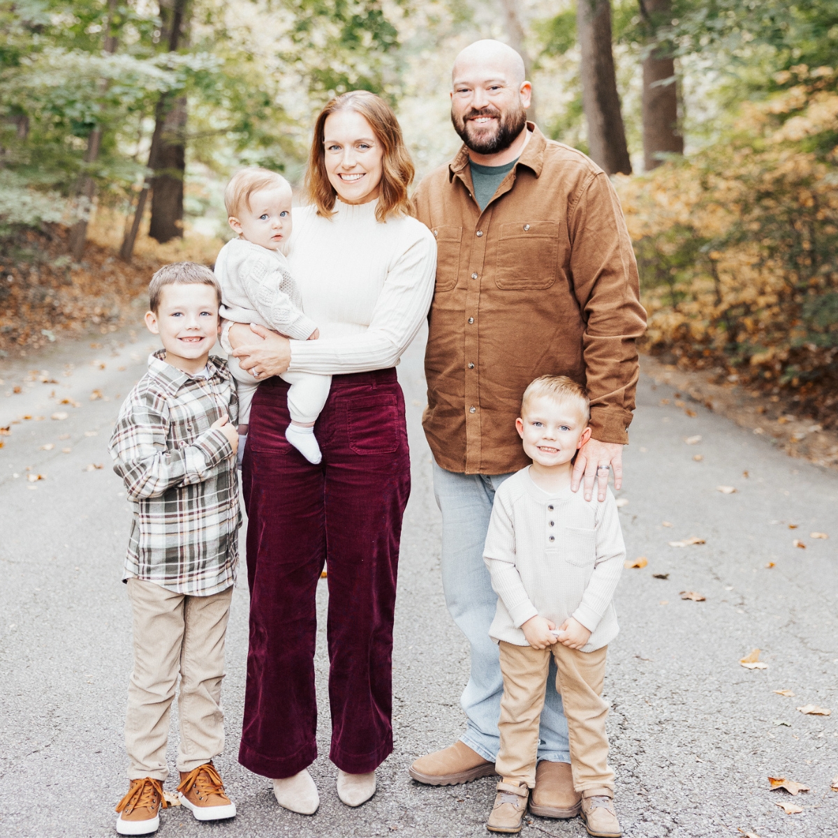 Smiling family of five standing on a road surrounded by trees with fall foliage.