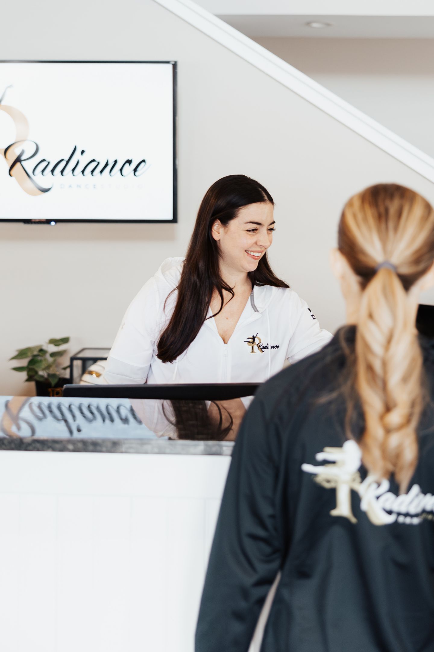 Smiling woman in white jacket behind reception desk at Radiance Dance Studio with a visitor in black jacket.