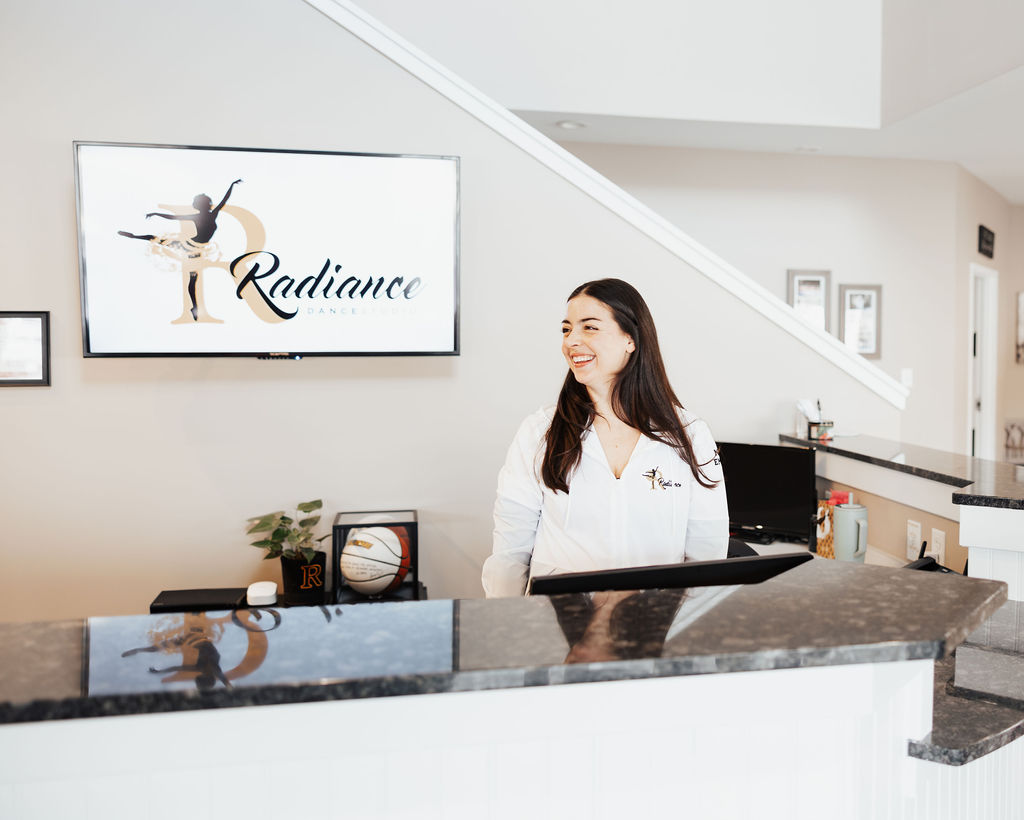Woman smiling behind a reception desk with a Radiance Dance logo on the wall behind her.