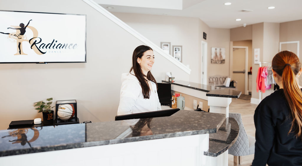 Woman smiles behind a reception desk at Radiance Dance Studio while another woman stands nearby.