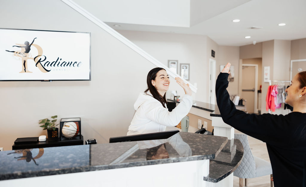 Two women smiling and high-fiving across a front desk in a bright dance studio reception area.