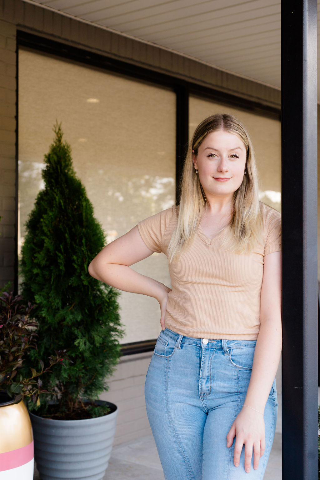 Young woman with blonde hair in beige shirt and jeans leaning against black post outside building.