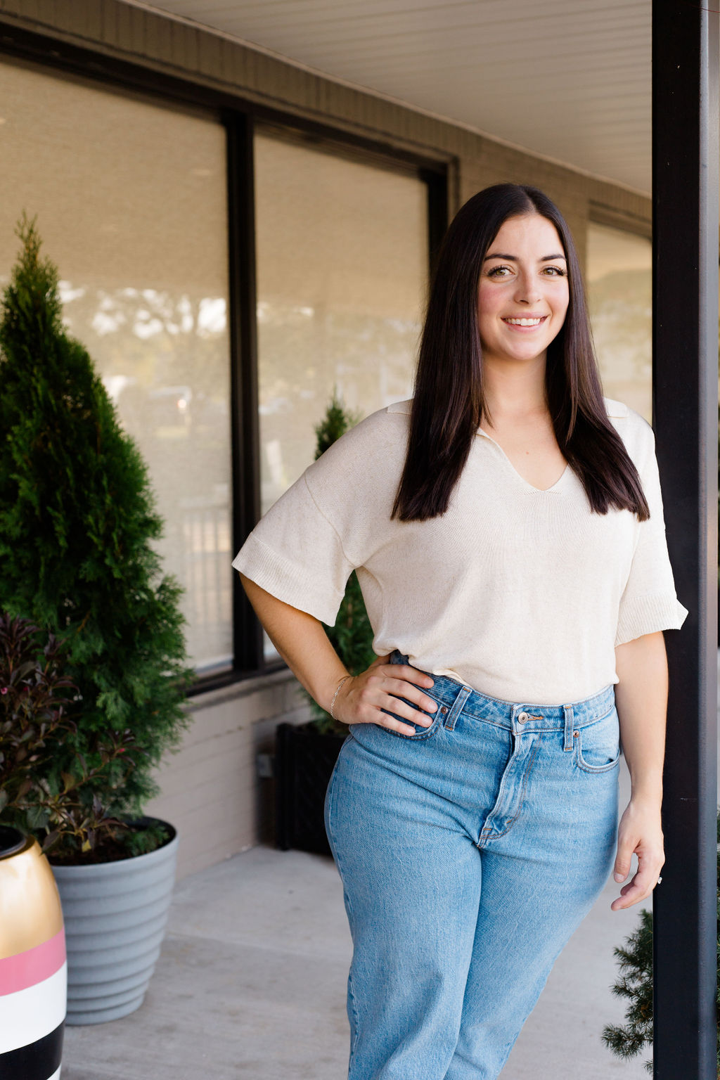 Smiling woman with long dark hair in white shirt and jeans stands with hand on hip outside building.