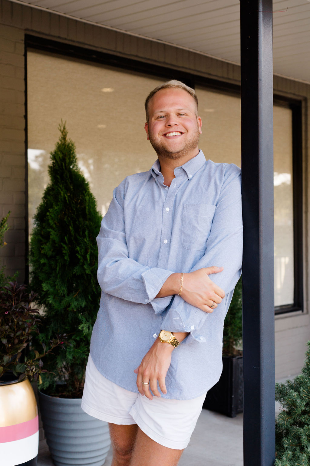 Smiling man in light blue shirt and white shorts standing outdoors by potted plants and window.