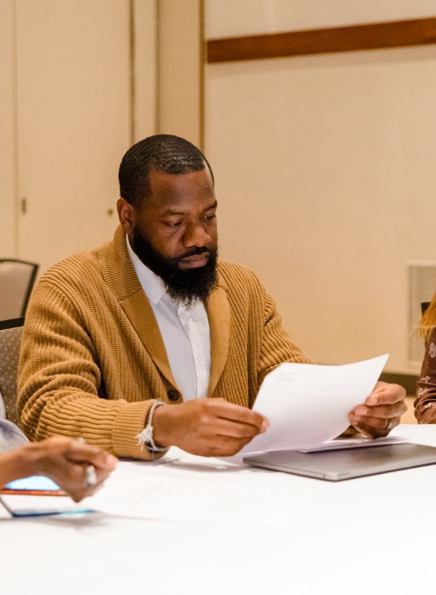 Man with beard wearing brown cardigan reading a document at a table with others in a meeting room.