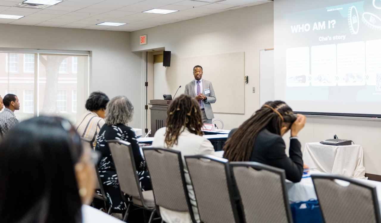 Man in suit presenting to seated diverse audience in a conference room with a projected slide.