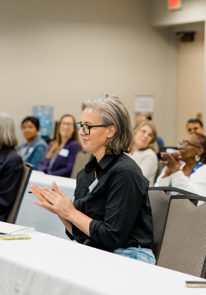 Woman with glasses clapping while sitting at a conference table with others in the background.