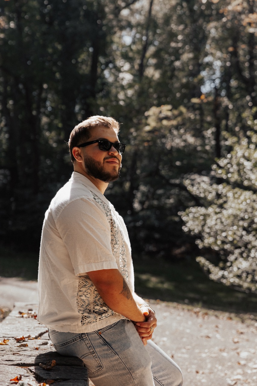 Man with beard and sunglasses wearing a white patterned shirt sitting outdoors on a stone ledge.