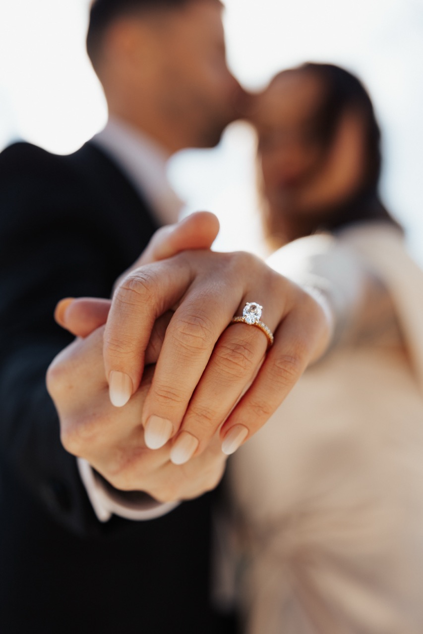 Close-up of a hand with an engagement ring held in front of a couple kissing.