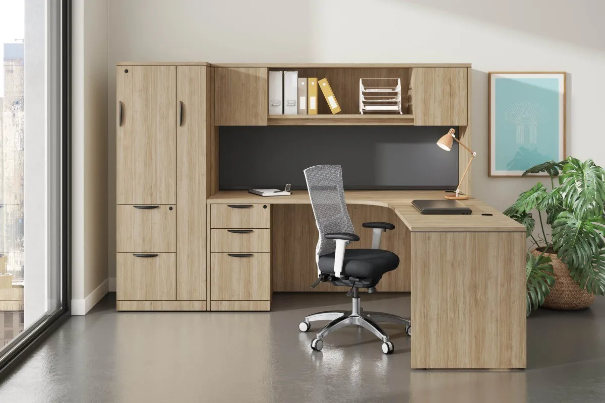 Modern office setup with a light wood L-shaped desk, ergonomic mesh office chair, desk lamp, binders, and a large plant near a window.