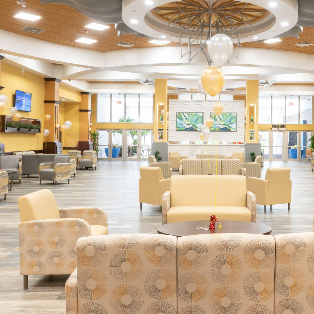 Bright and spacious lobby area by Lesro, with patterned and plain beige sofas, round tables, yellow and white balloons, and colorful plant paintings on the far wall.