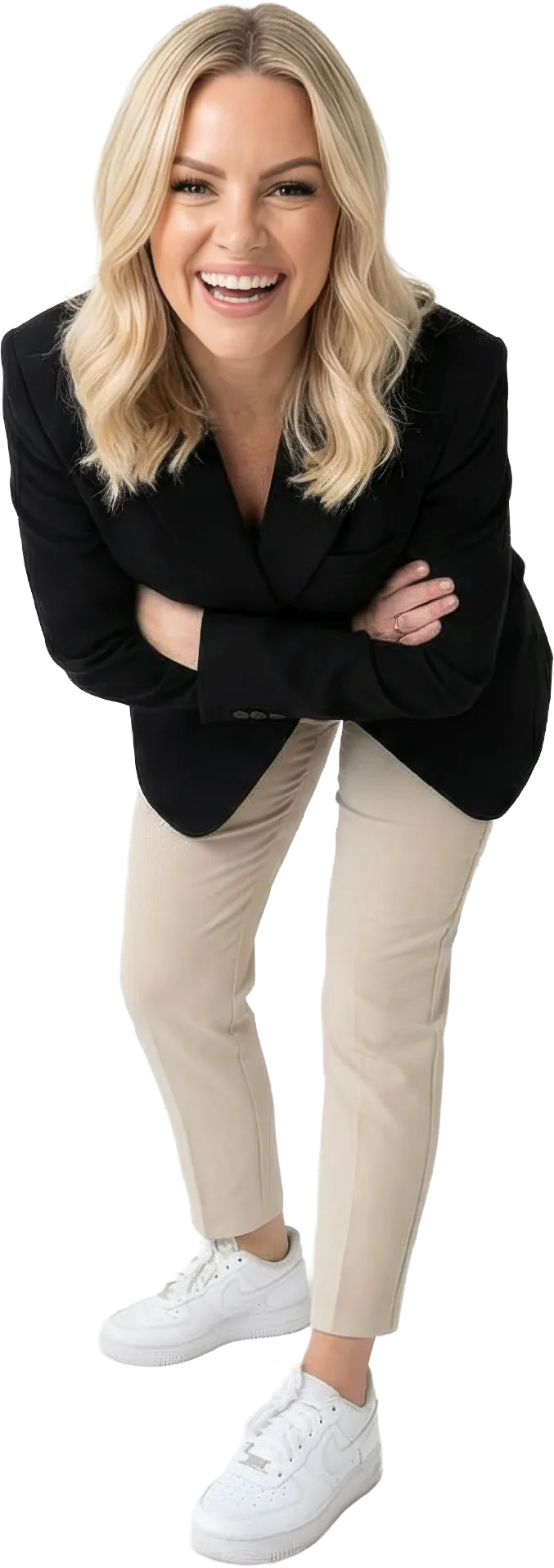 Smiling blonde woman wearing black blazer, beige pants, and white sneakers, leaning forward with arms crossed.