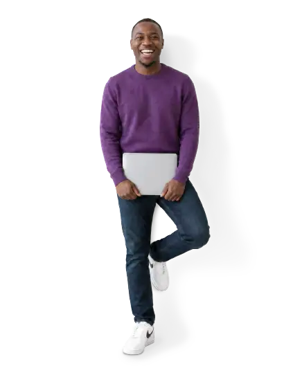 Smiling man in purple sweater holding a laptop, standing with one foot raised and wearing jeans and white sneakers.