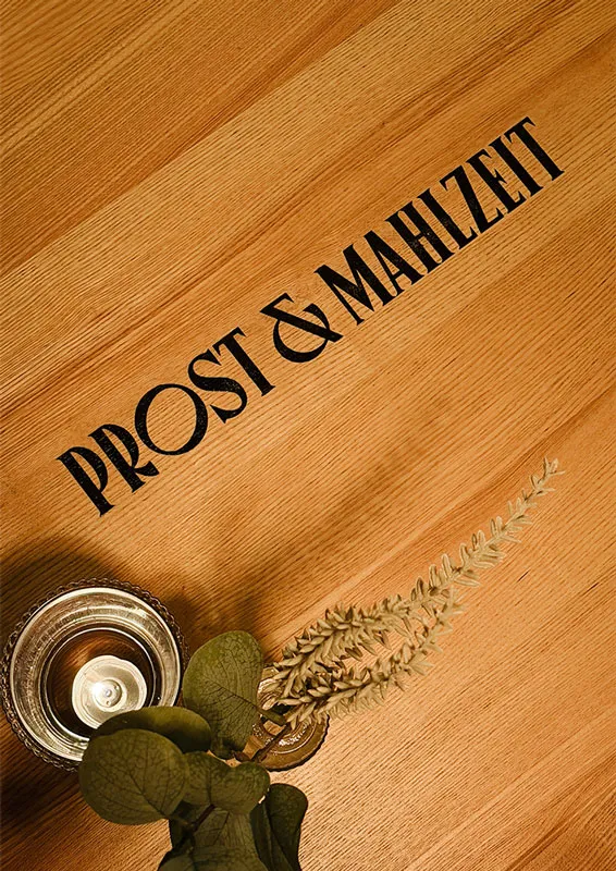 Prost & Mahlzeit steht auf einem Holztisch geschreiben, daneben ein Teelicht und ein Blumengesteck