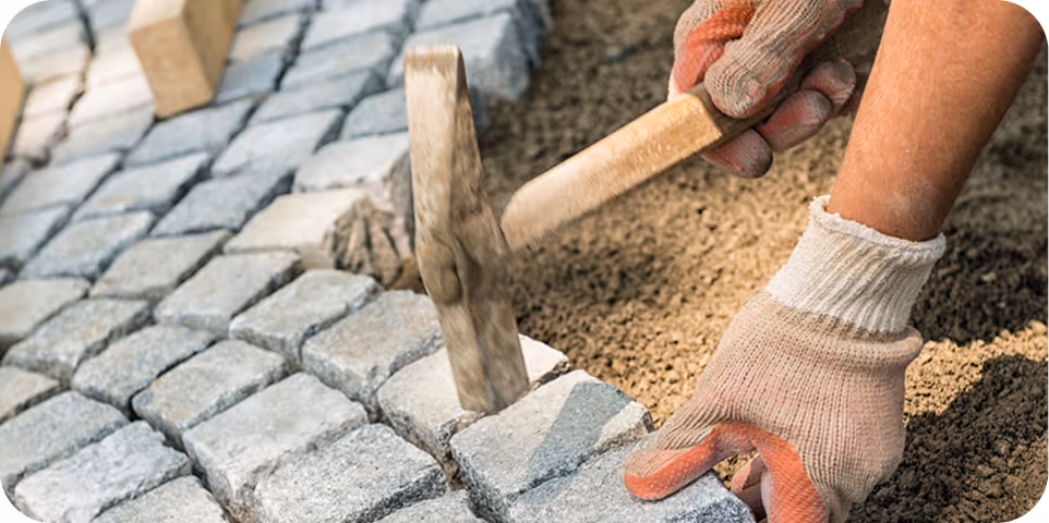 Nahaufnahme einer Person mit Handschuhen, die Kopfsteinpflaster mit Hammer und Sand verlegt.