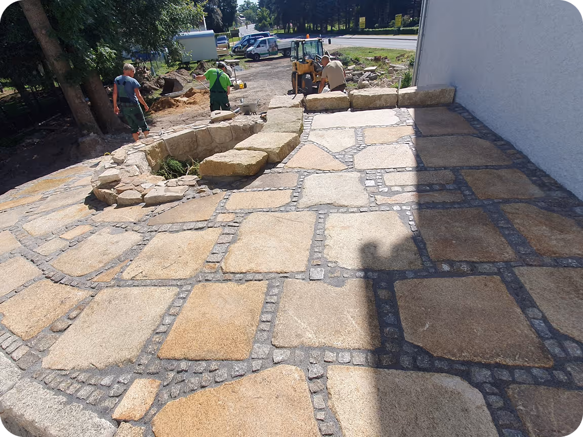 Drei Bauarbeiter verlegen große rechteckige Natursteinplatten zu einer Terrasse neben einem weißen Haus, im Hintergrund Baufahrzeuge und Bäume.