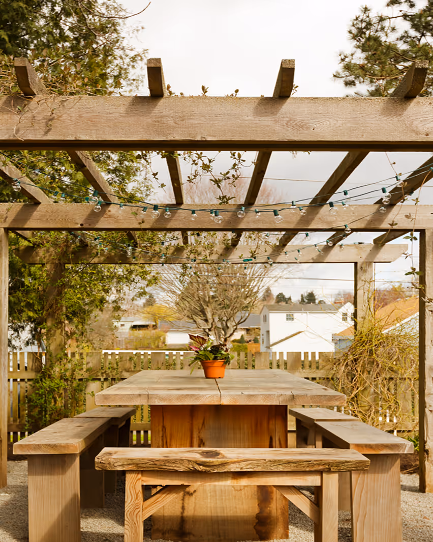 Holztisch mit Bänken unter einer Pergola mit Lichterketten und einem Blumentopf im Garten.