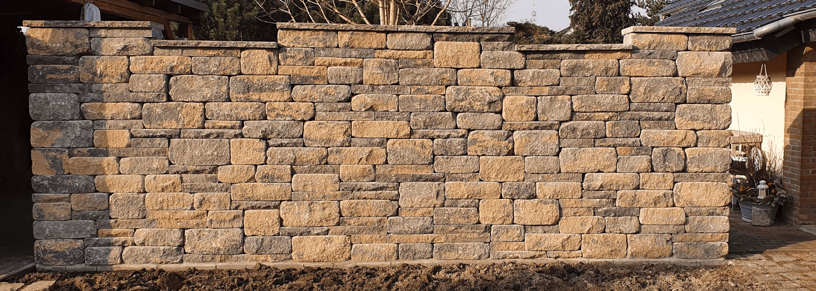 Natursteinmauer mit rechteckigen Blocksteinen in zwei Farbtönen, gebaut vor zwei Gebäuden im Hof.