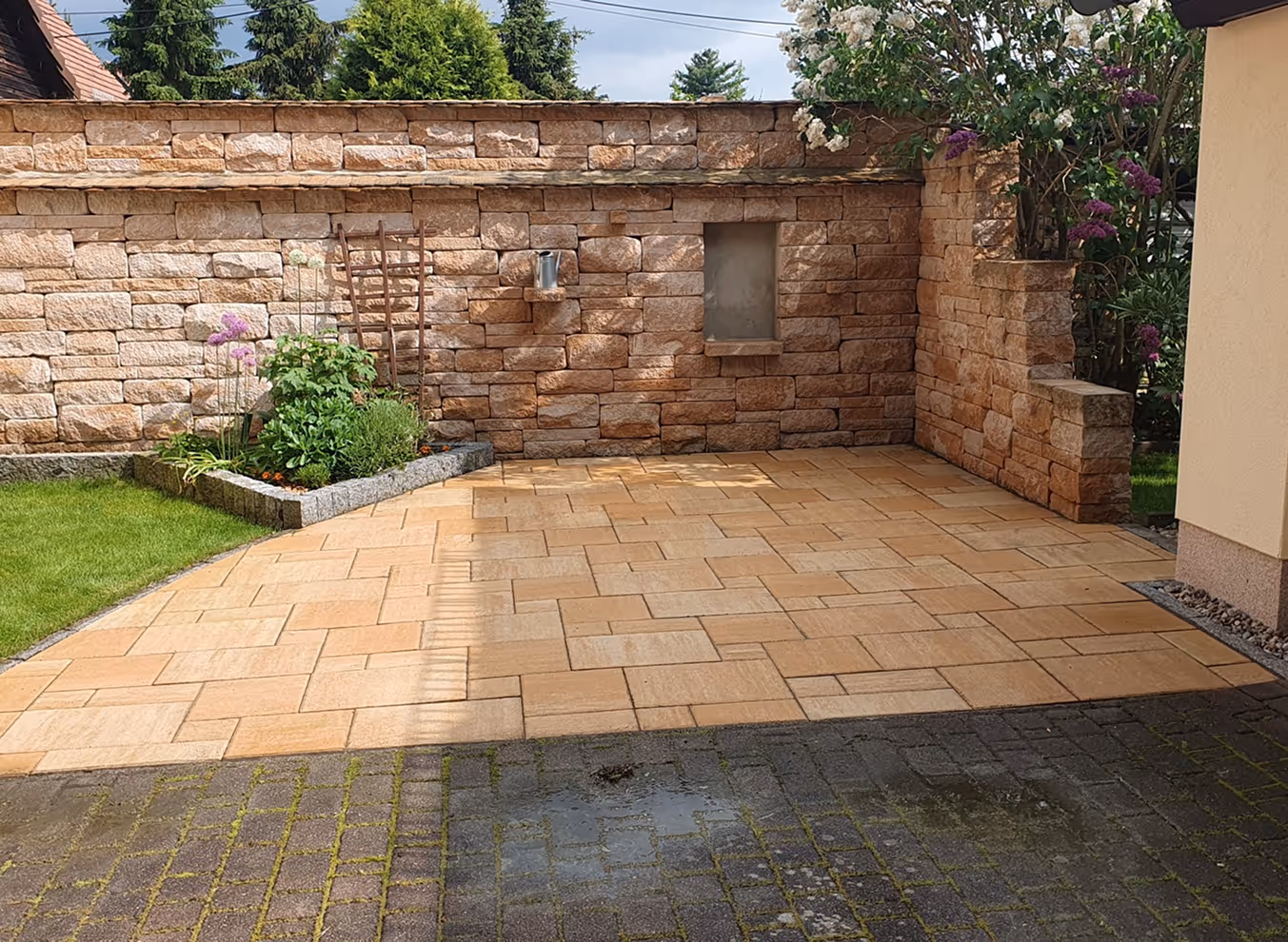 Gepflasterte Terrasse mit hellbraunen Steinen vor einer Mauer aus großen Natursteinen und einem kleinen Blumenbeet mit grünen Pflanzen und lila Blüten.