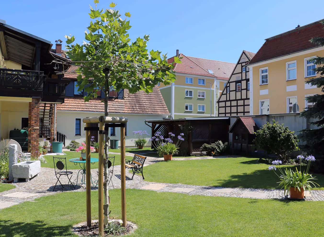 Gut gepflegter Garten mit grüner Wiese, jungen Bäumen, Blumen in Töpfen, Gartenmöbeln und historischer Fachwerkhaus- und Wohnhauskulisse bei blauem Himmel.