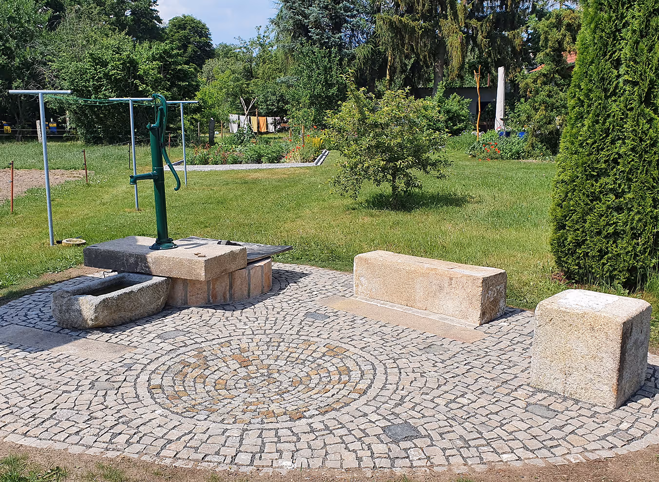 Grüne Handpumpe auf gepflastertem Platz mit Steinbank und Natursteinbrunnen, umgeben von Gras und Bäumen.