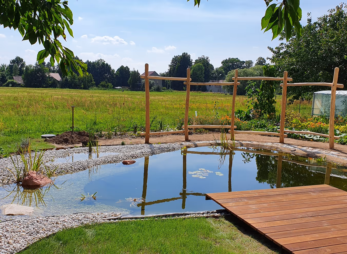 Natürlicher Gartenteich mit Holzsteg, Kiesumrandung und umgebender grüner Wiese unter blauem Himmel.