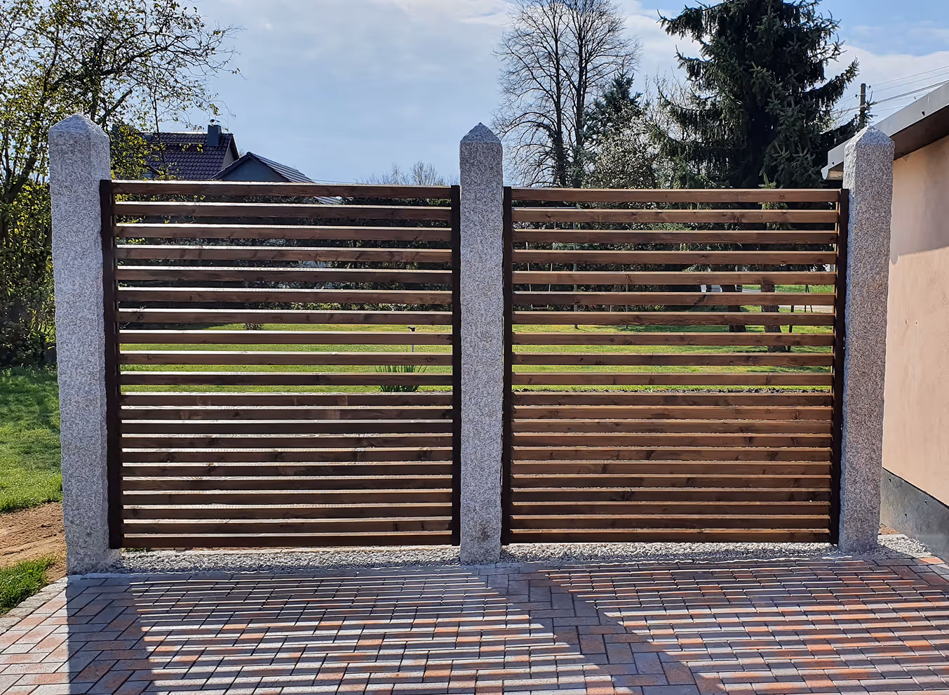 Moderner Holzzaun mit horizontalen Latten und grauen steinernen Pfosten vor einem gepflasterten Hof.
