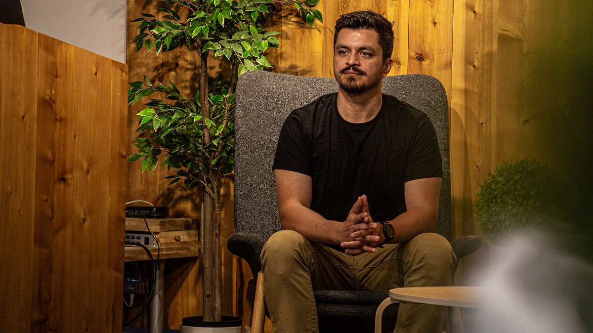 Man with dark hair and beard sitting on a gray armchair in a wood-paneled room with green plants.