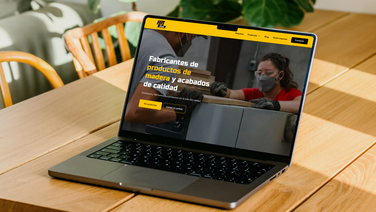 Laptop on wooden table displaying HR Wood website homepage with a banner showing two people working with wood and text about quality wood products.