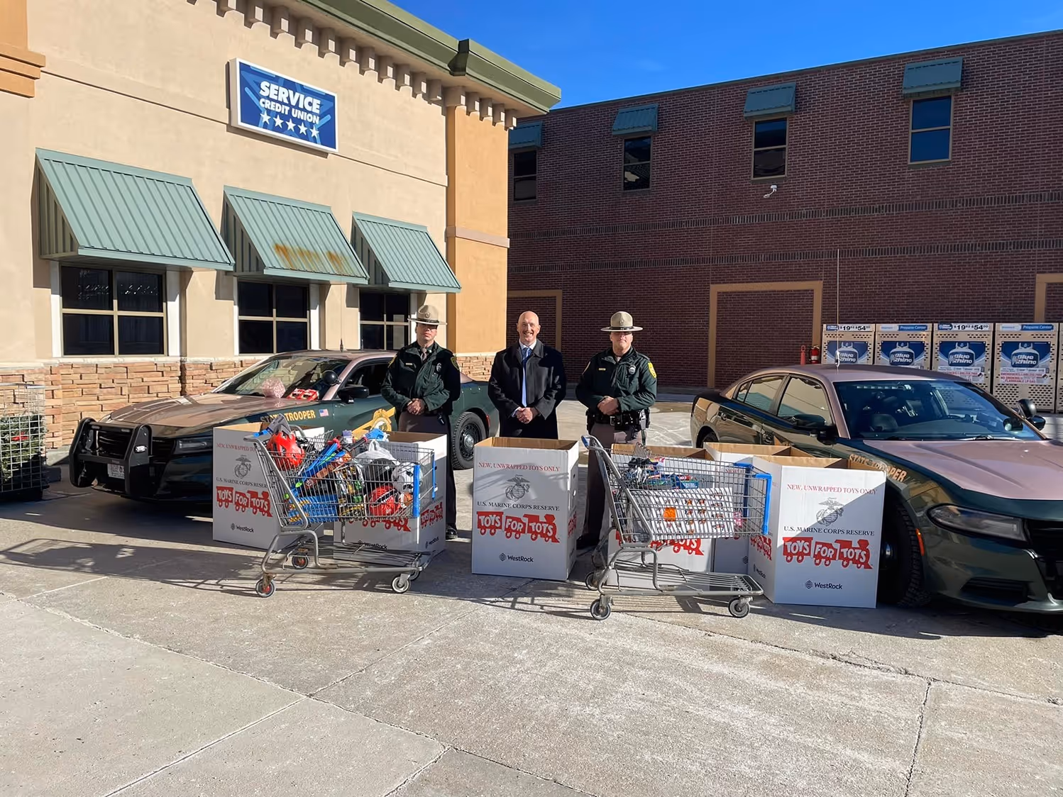 Two state troopers and a man in suit standing between two police cars with shopping carts and boxes filled with Toys for Tots donations outside a credit union building.